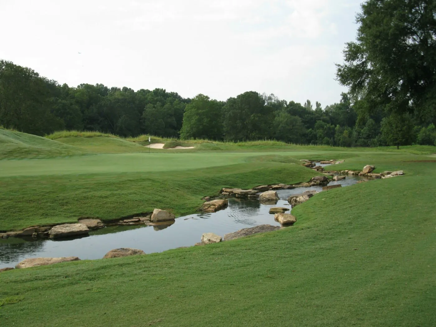 A green and its bunker beyond a rock-lined stream, with a wooded ridge behind.
