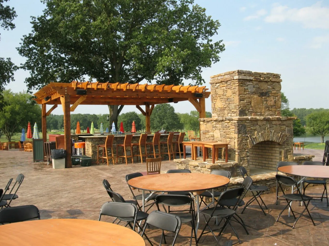 A covered pergola and tall stone fireplace on a paved patio set with tables and chairs, umbrellas closed.