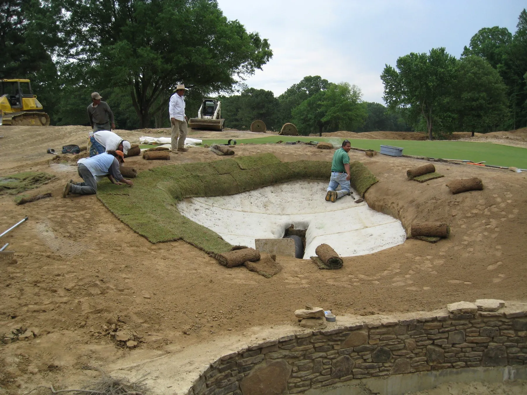 Crew members laying sod around a newly lined bunker, rolls of turf stacked on the bank and a stone wall below.