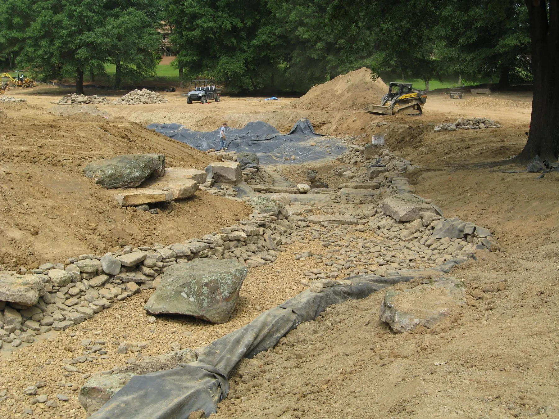A streambed under construction — stacked rock along the banks, gravel and liner in the channel, and a skid steer working at the top.