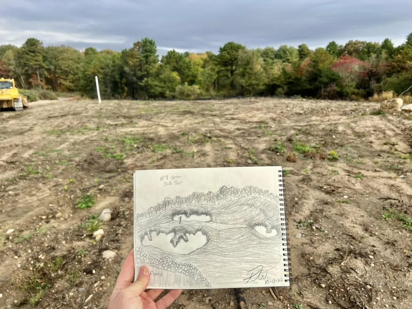 A hand holds up a pencil sketch of a green and its bunkers against the bare graded ground where it will be built.